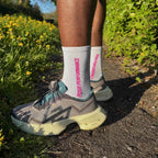 Person wearing grey running shoes and white performance running socks with pink text, standing on a path with greenery.