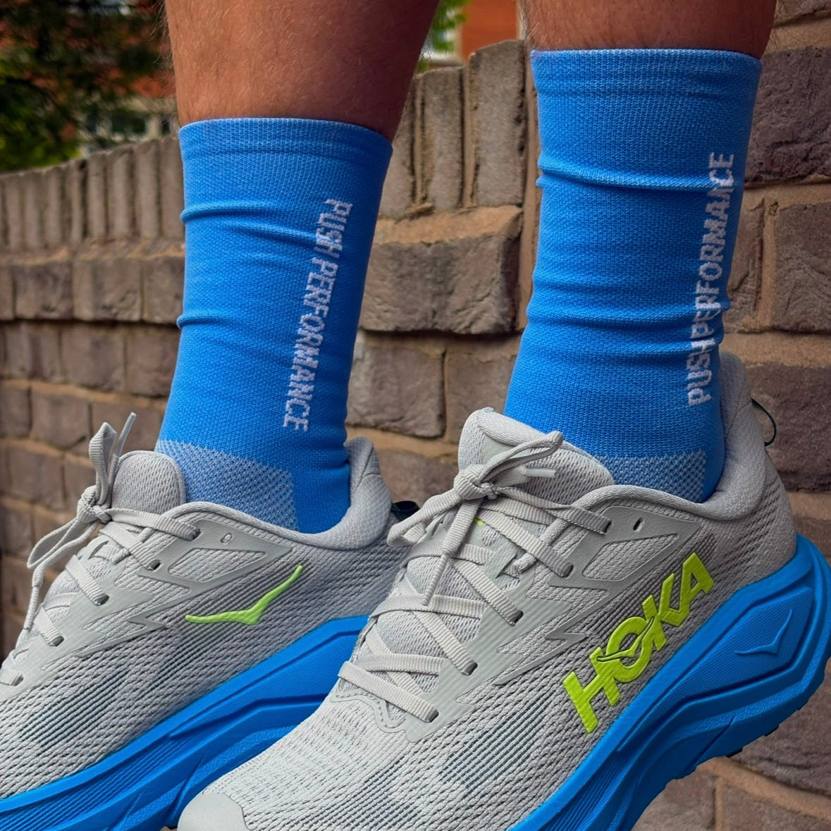 A close-up of a person's legs wearing grey and blue Hoka running shoes with bright blue crew running socks featuring white 'PUSH PERFORMANCE' text, set against a brick wall background.