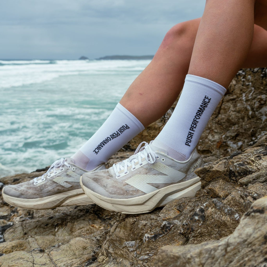 Person wearing white socks with text and sneakers sitting on rocks by the ocean.