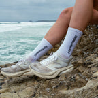 Person wearing white socks with text and sneakers sitting on rocks by the ocean.