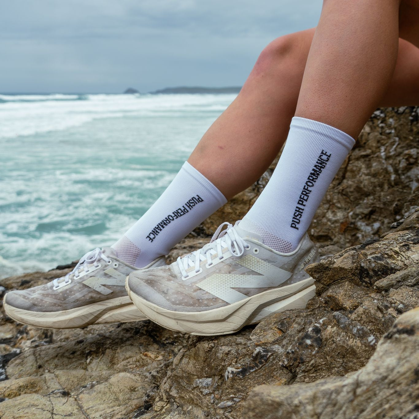 Person wearing white socks with text and sneakers sitting on rocks by the ocean.