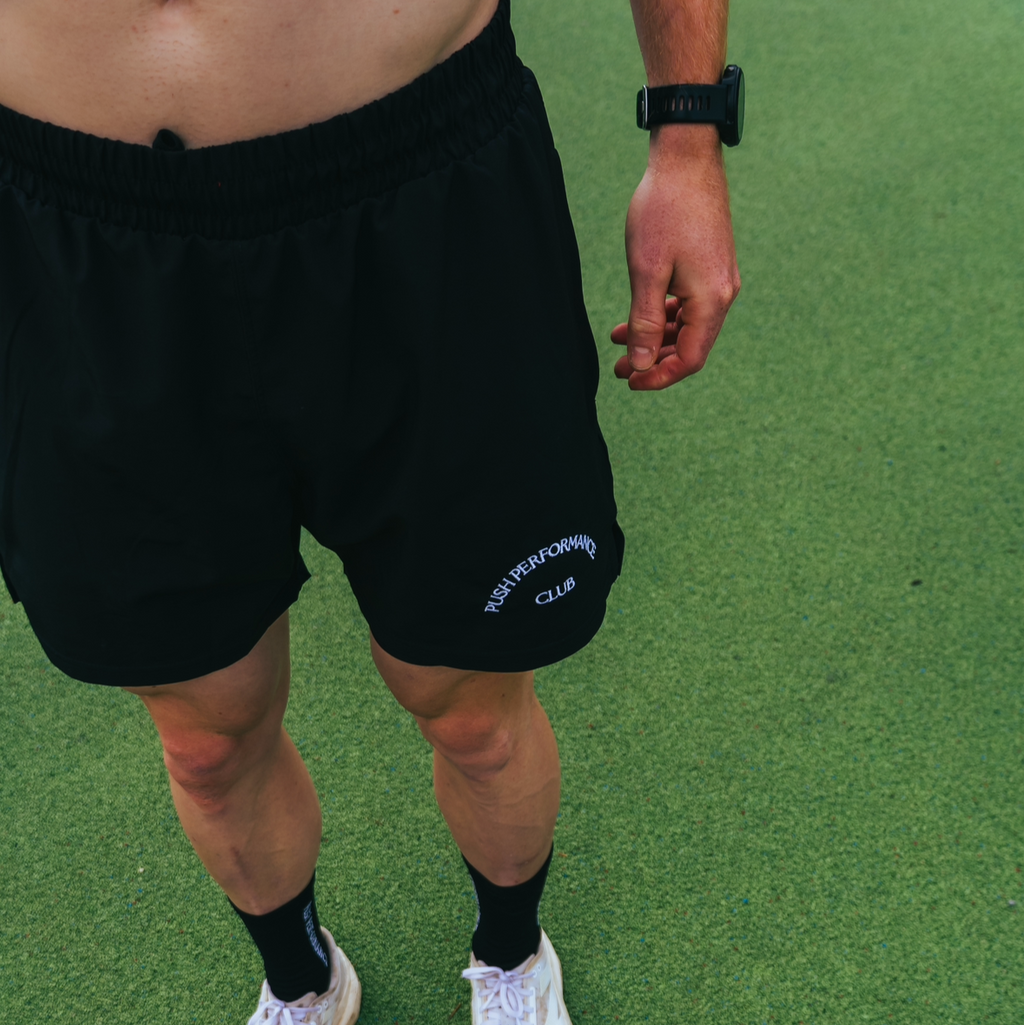 Person wearing PUSH PERFORMANCE  black shorts with a logo on a green grassy field.